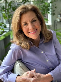 Smiling woman with shoulder-length hair in a purple shirt, sitting indoors with leafy plants in the background.