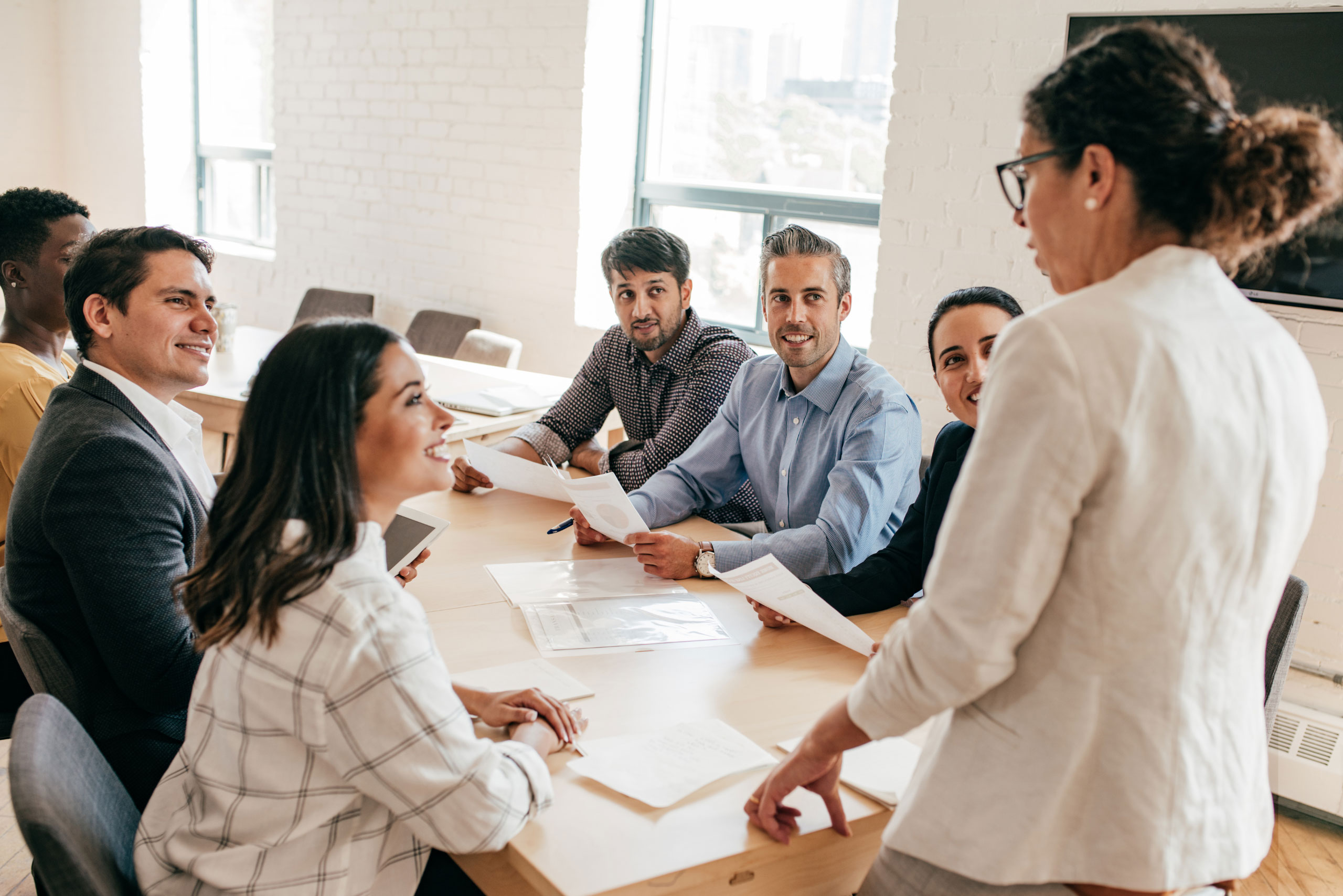 A group of seven people in a meeting around a table, smiling and looking at a woman standing and talking.