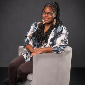 A woman with braided hair, glasses, and a patterned blouse sits on a gray chair against a dark background, smiling.