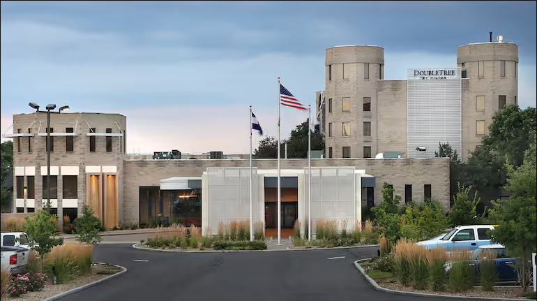 Three-story DoubleTree hotel with flags in front and a round driveway entrance, set against a cloudy sky backdrop.