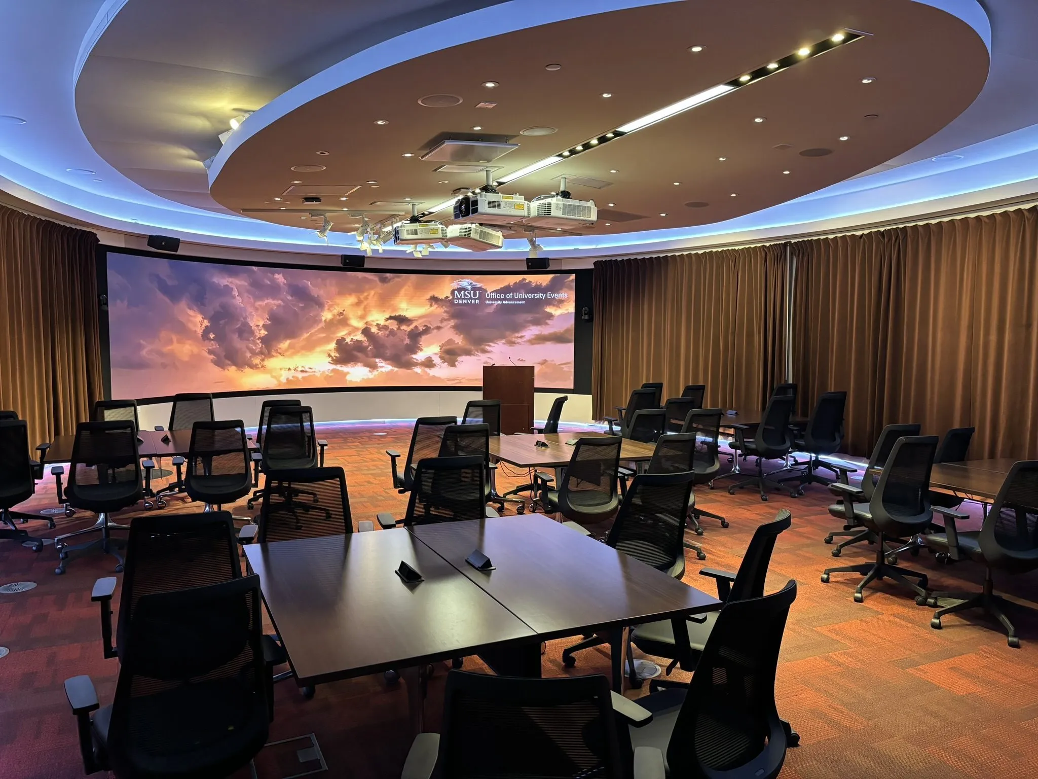 Modern conference room with multiple tables and chairs, featuring a large curved screen displaying a scenic sky image.