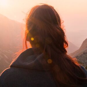 Back view of person with long hair in a braid, standing on a mountainside, watching the sunrise over distant peaks.