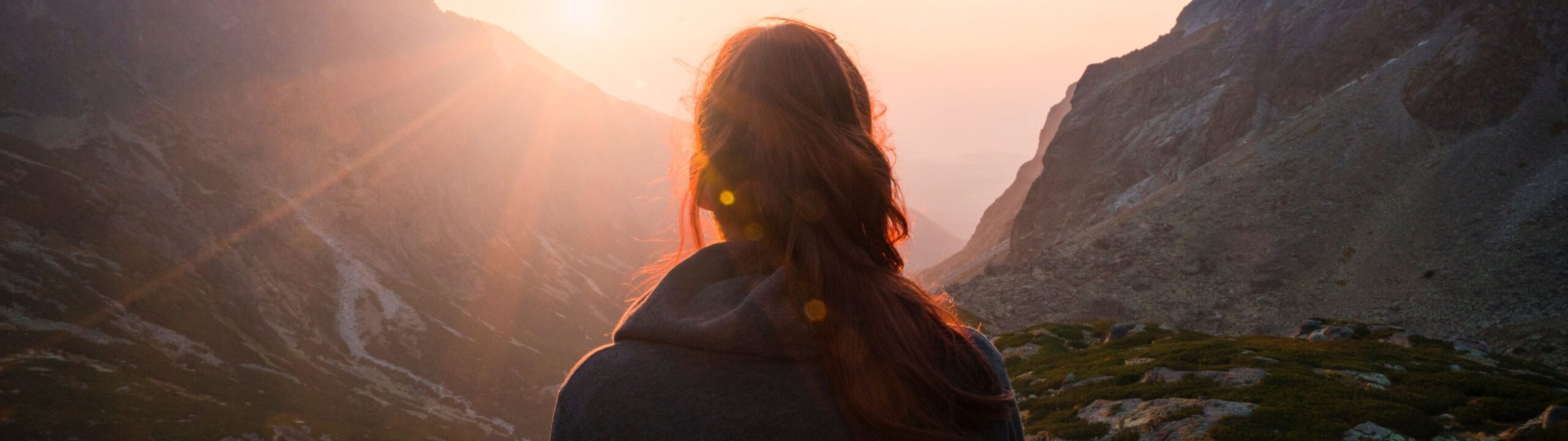 Back view of person with long hair in a braid, standing on a mountainside, watching the sunrise over distant peaks.