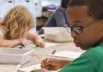 Children are seated at a classroom table, focused on writing and drawing activities.