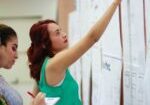 Three women stand and examine papers on a bulletin board; one points while another takes notes.